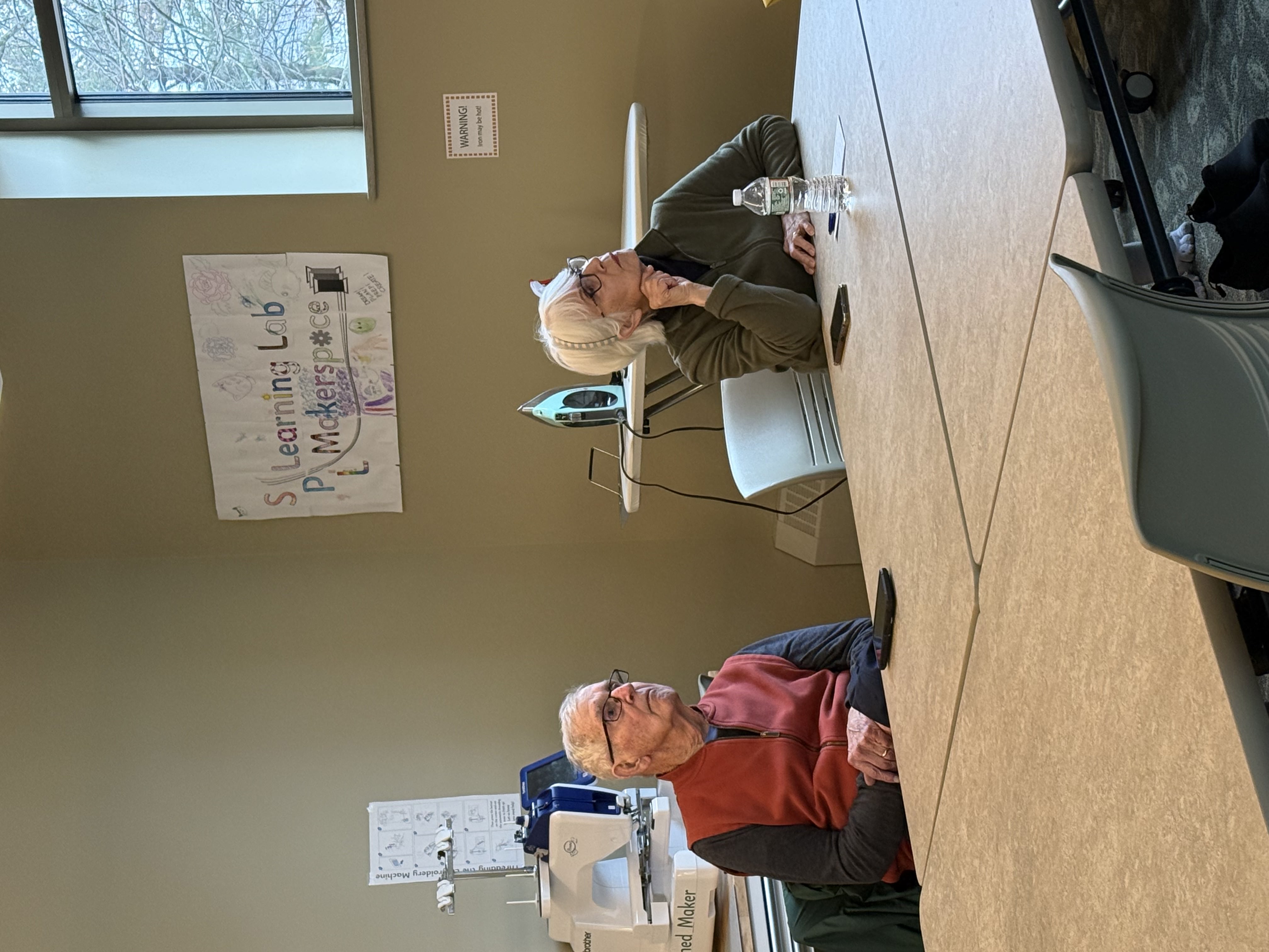 Two older adults at a workshop table getting one-on-one tech help